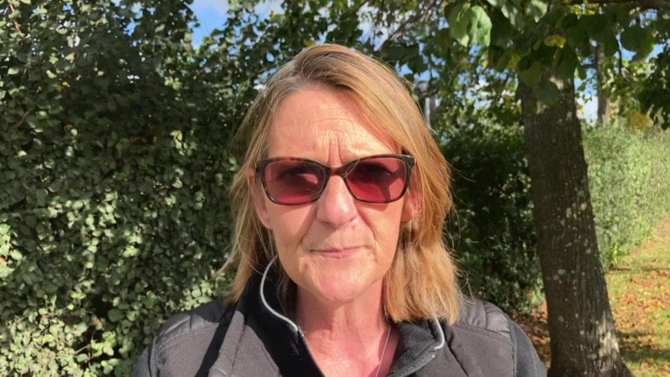 Nicola Sharpe has sunglasses on and is standing in front of a green shrub.
