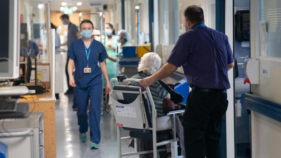 An older person with white hair is pushed in a hospital chair by a man in blue medical uniform in a hospital corridor