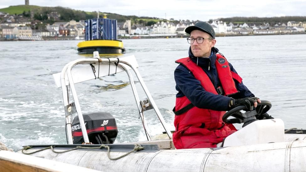 Man in a red flotation suit operates a small inflatable workboat in a harbour, with a floating marine device and waterfront buildings visible in the background.