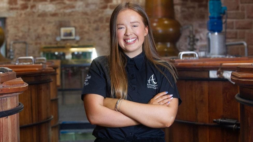 A young woman with long brown hair in a black Annandale Distillery T-shirt stands with her arms folded, smiling. Behind her are the wooden containers and pipes of a whisky distillery.