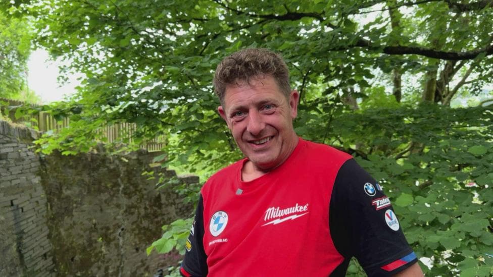 Man with curly brown hair and smiling at the camera is wearing a red and black t-shirt with a backdrop of leafy trees.