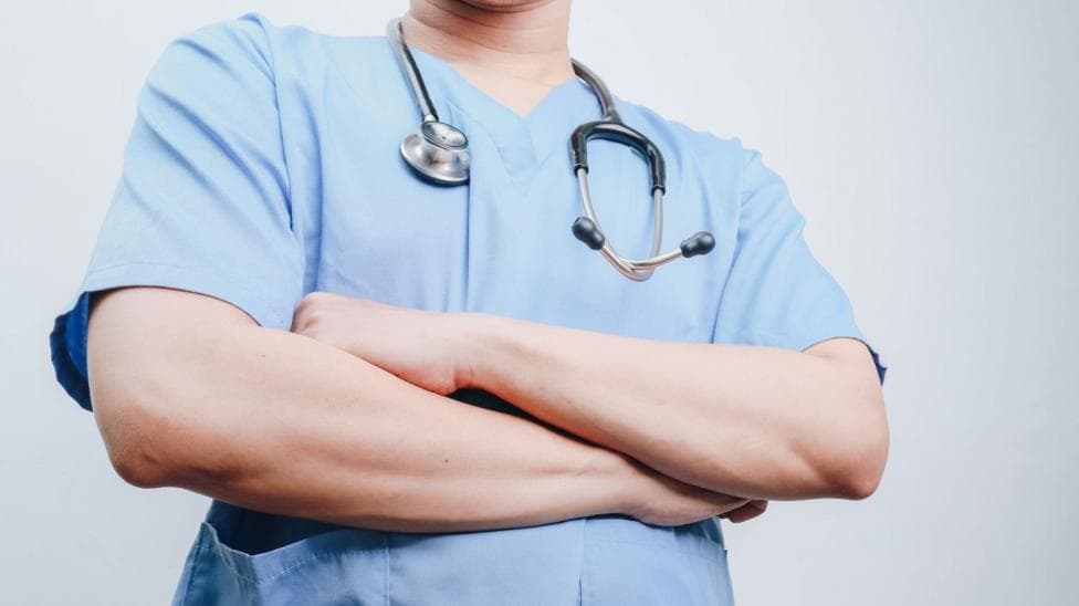 A person wearing medical scrubs has a stethoscope around their neck and is folding their arms.