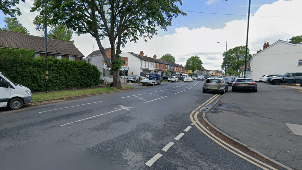 A generic photo of Shaftmoor Lane at a junction with another street, part of which is visible in the bottom right of the image. There are houses and some trees to the left of Shaftmoor Lane on the photo.