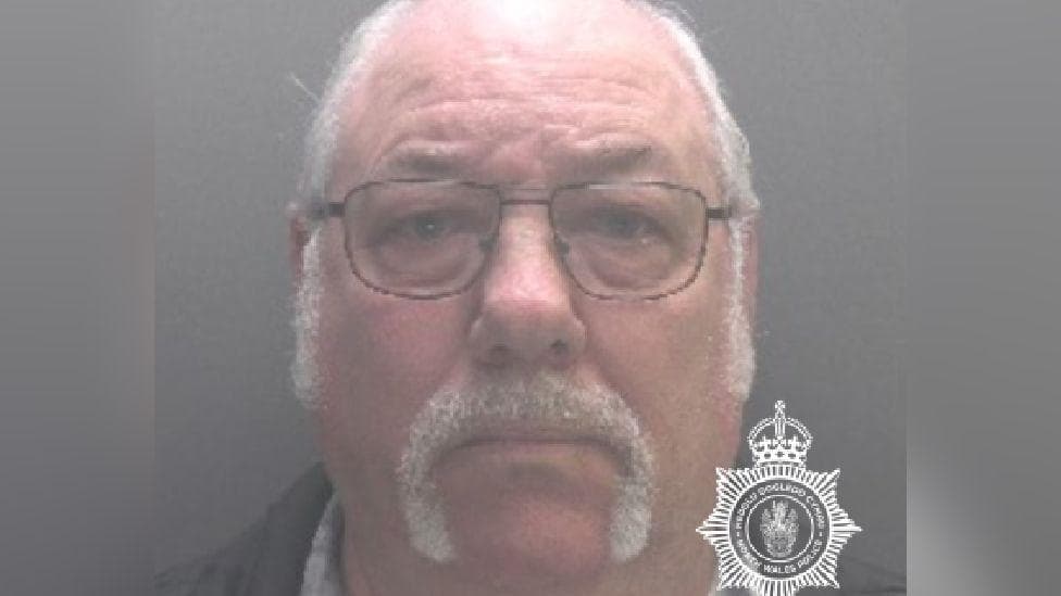 Mugshot of Arthur Hughes. He has white short hair and a white moustache which reaches his chin. He wears glasses with dark frames. He is looking at the camera and not smiling.