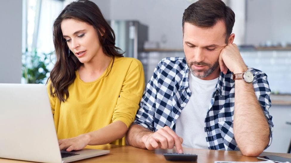 Couple sit at a kitchen table with the woman in a yellow jumper looking at a laptop and the man in a blue and white shirt pressing numbers on a calculator.