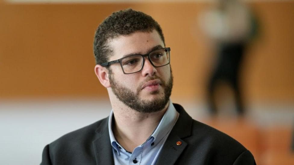 A man with brown hair and beard, wearing black-rimmed glasses and a dark suit