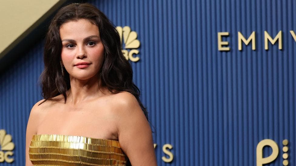 Selena Gomez at the 2026 Emmy Awards, wearing a gold strapless gown, and posing against a dark blue background