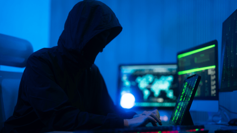 A person wearing a hooded coat is typing on a computer. They have multiple screens open and appear to be doing something covert. The room is illuminated with a dark blue light.