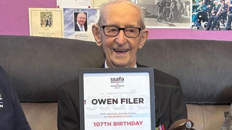 A 107-year-old man smiling at the camera and holding a framed certificate