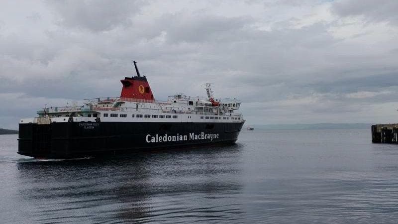 MV Caledonian Isles sailing across a calm harbour toward a pier on the right, beneath a cloudy grey sky. The black-and-white vessel is reflected in the still water, with distant coastline visible on the horizon.