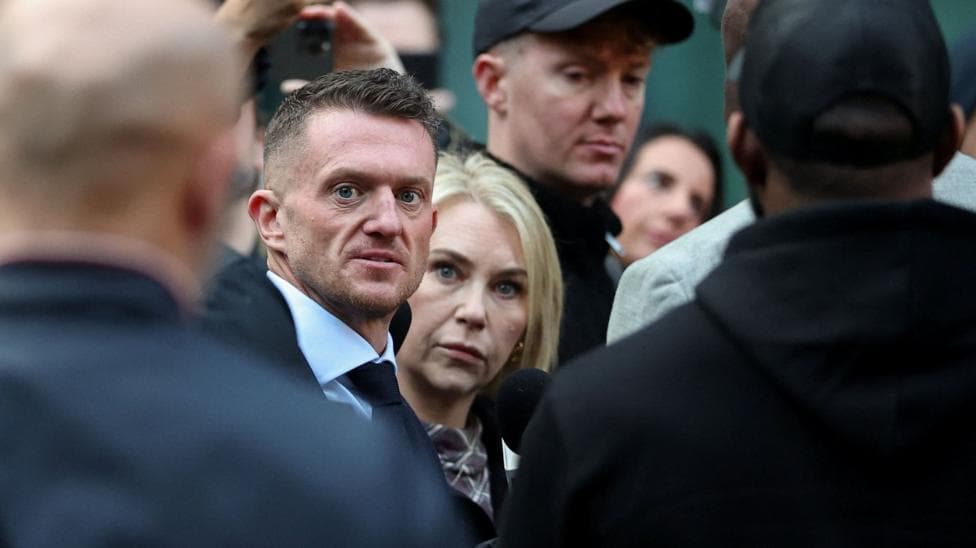 Tommy Robinson stares at the camera as he walks into court, surrounded by a close crowd and security officers. He is wearing a white shirt and black tie. Next to him is a woman with blonde hair and blue eyes, who also stares right at the camera.