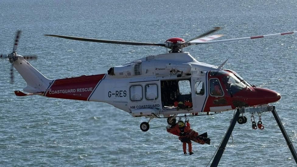 A man being winched to safety by a coastguard helicopter coloured red and white. They are flying above the water.
