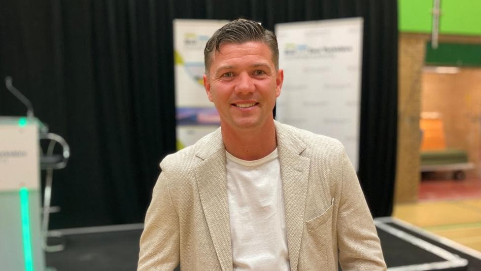 Luke Campbell standing by a stage on election night. He has short dark hair and is wearing a cream jacket and white T-shirt