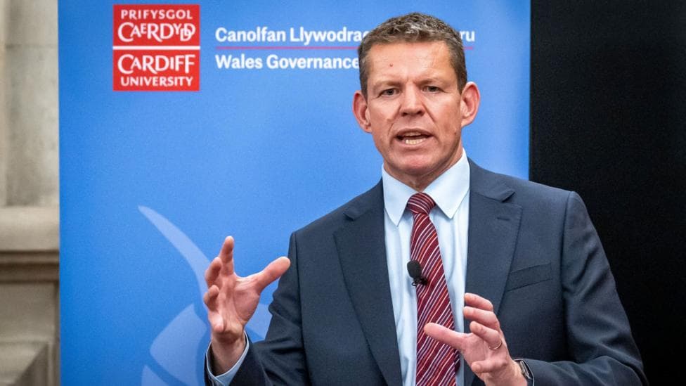 The first minister Rhun ap Iorwerth stood in front of a banner advertising Cardiff University's Welsh governance centre. The banner is blue and has a red Cardiff University logo. Rhun wears a grey suit, white shirt, and red tie with diagonal stripes.