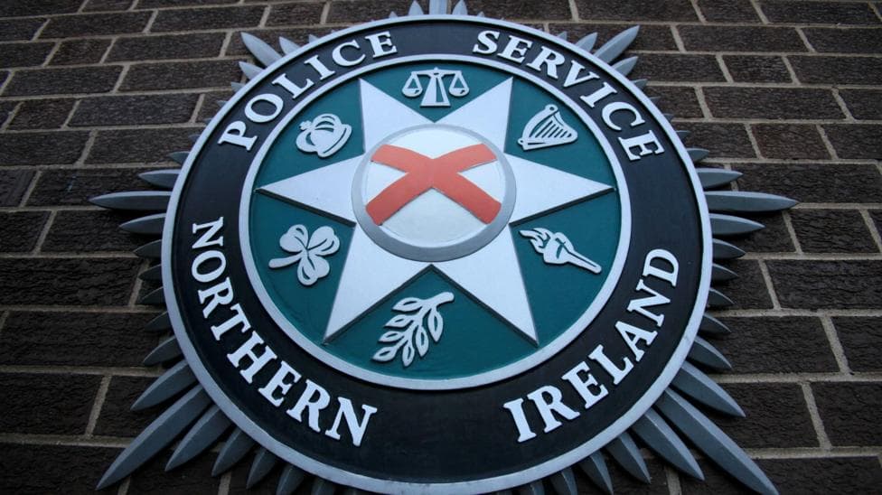 The PSNI emblem on a brick wall. Saint Patrick's satire is in the centre of a star shape, surrounded by scales, a harp, a torch, a leaf, a shamrock and a crown.