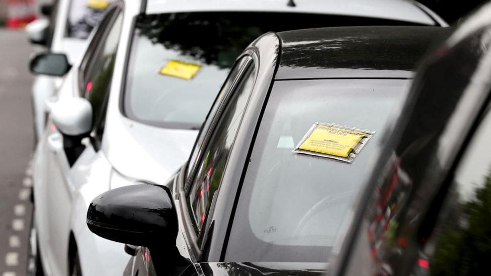 A generic-looking image shows a row of cars seen front-on with yellow and black Penalty Charge Notices affixed to their windscreens, on a road near Gatwick Airport in 2017.