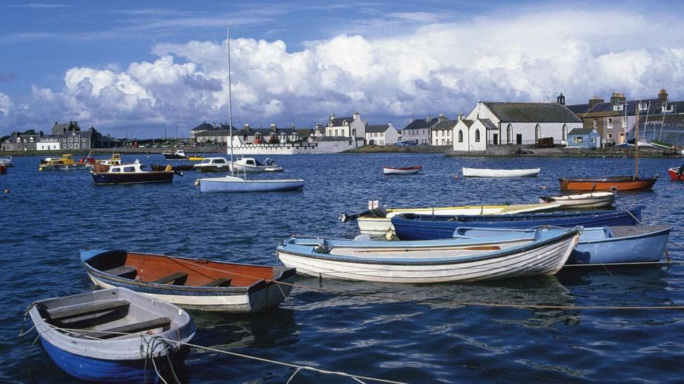 Some small boats tied up at the shore with the the fishing village of Isle of Whithorn in the background with clouds building up over blue skies
