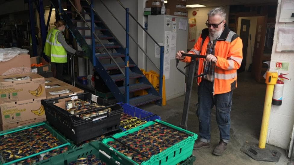 Inside FareShare South West's distribution hub, a volunteer wearing a high visibility orange jacket moves a green crate with a manual pallet truck. Crates of food and boxes of bananas are stored nearby, while two other volunteers work in the background.