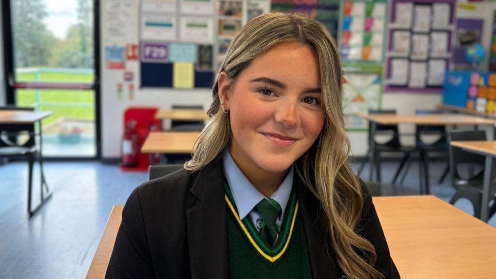 A female student wearing a school uniform sits at a desk in a classroom, facing the camera. The uniform includes a dark blazer, light blue shirt, green tie and green sweater with yellow trim. The classroom contains rows of desks and chairs, colourful educational displays on the walls, and a glass door with a view of greenery outside.