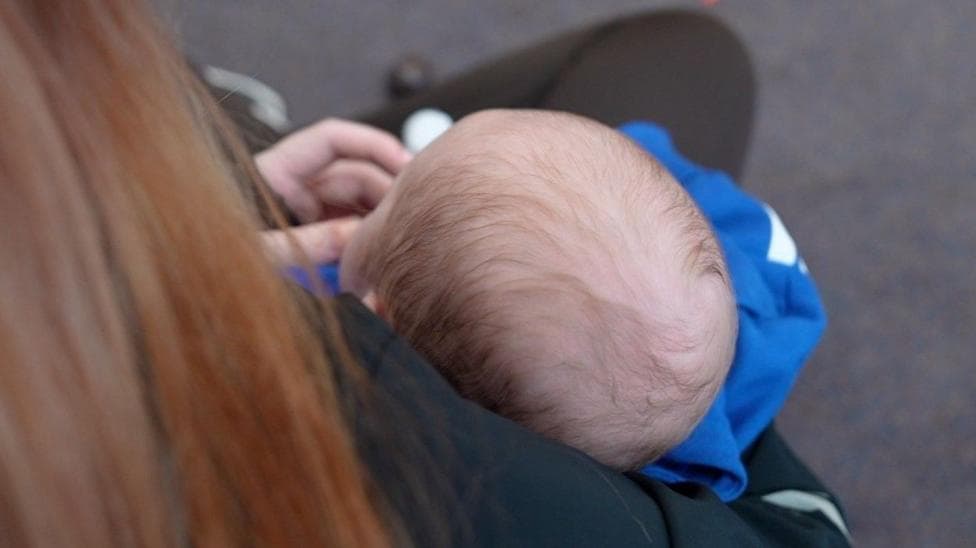 The picture is taken from behind a young mum with light brown long blonde hair. We can't see her face but we can see over her shoulder, she is holding a baby in her arms. The young baby is wearing blue and has dark hair.