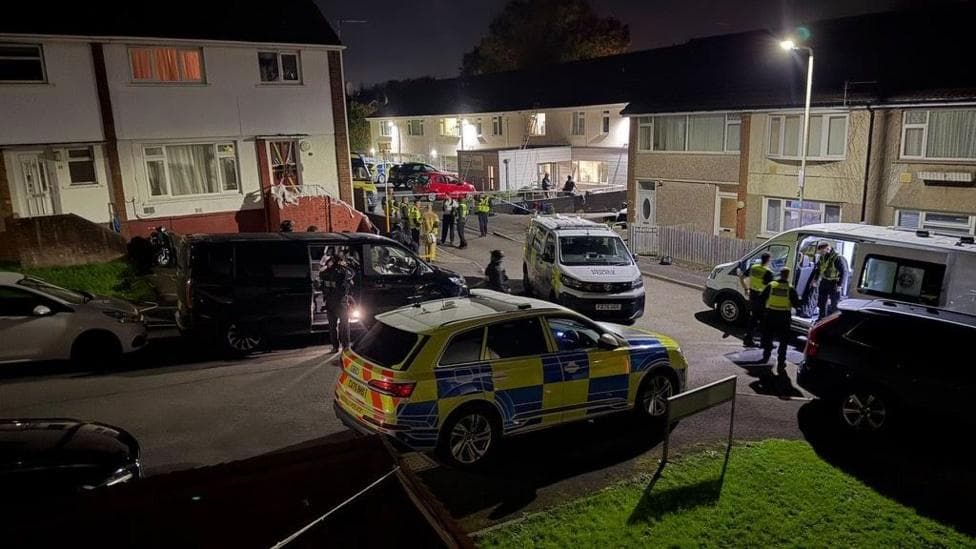A road on a housing estate and with loads of cars along with police cars and vans and police officers.