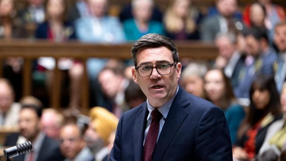 Andy Burnham is wearing glasses, a dark-coloured suit, red tie and white shirt. He has short black hair. Burnham is stood up and speaking in the House of Commons. Behind him are several MPs, who are sat down and out of focus.