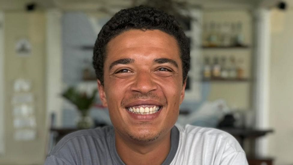 Michael Ashton is wearing a T-shirt that is half grey and half white. He has short black hair and is smiling. He is sitting in what appears to be a kitchen.