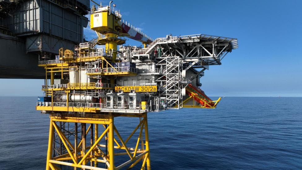 A mostly yellow-painted gas platform and allied grey rig stand in calm blue seas with a clear blue sky behind them. It is sunny.