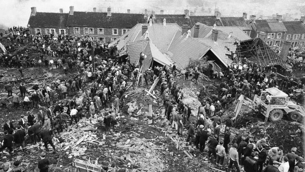 A black and white photo of Pantglas Junior School, collapsed under the weight of mud, and villagers and emergency services helping to free trapped children and adults.