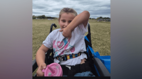 Remi making a thumbs up sign in her wheelchair as she wears a colourful T-shirt with a pink her