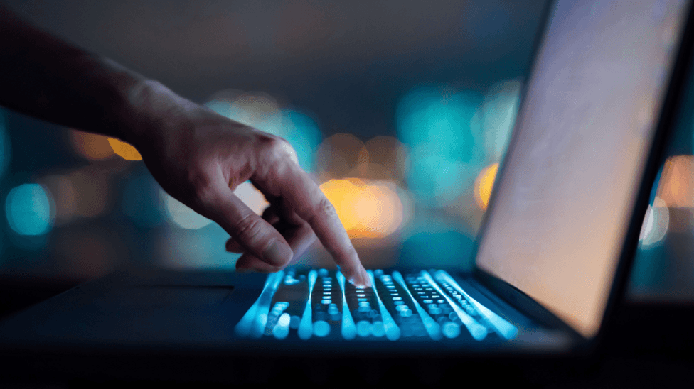 A close-up of a woman's hand typing on a computer keyboard in the dark against colourful lights in background. The keyboard is lit up.