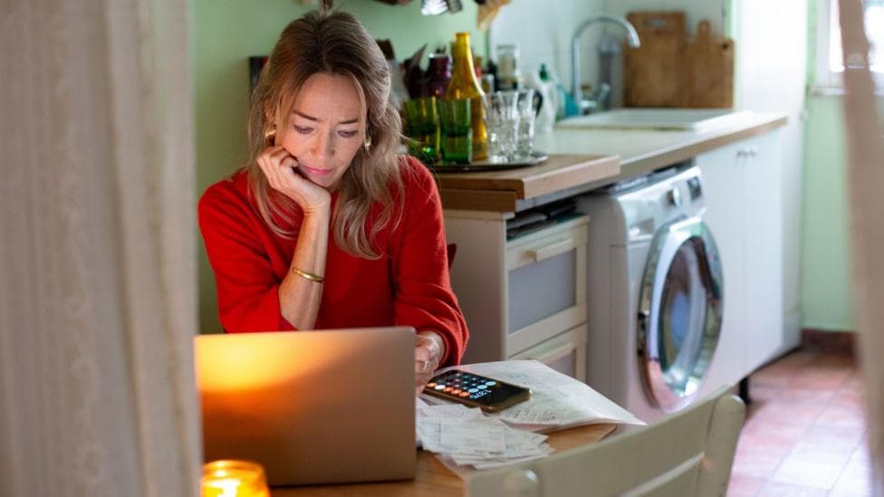 A woman sits looking at a laptop in a kitchen - stock shot.