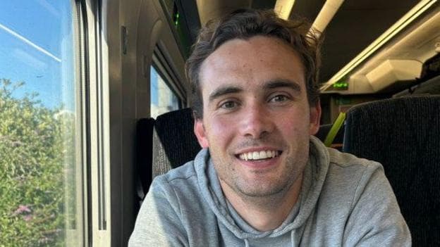 Tufnell, a man with brown hair, sat in a train carriage, wearing a grey hoodie and smiling as he looks at the camera.
