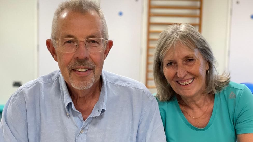 Steve and Angela Bucksey sit side-by-side smiling - Steve has grey hair and is wearing a light blue shirt with a collar, and Angela has shoulder-length greyish hair and wears a green round-necked T-shirt and a necklace