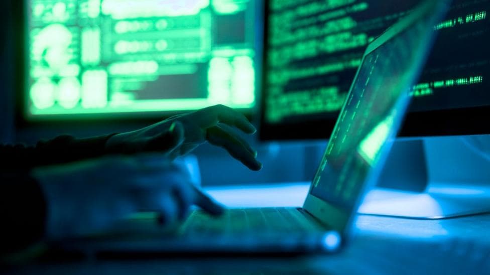 A pair of hands at a laptop with two computer screens in the background - both with green lines of code on a black backdrop