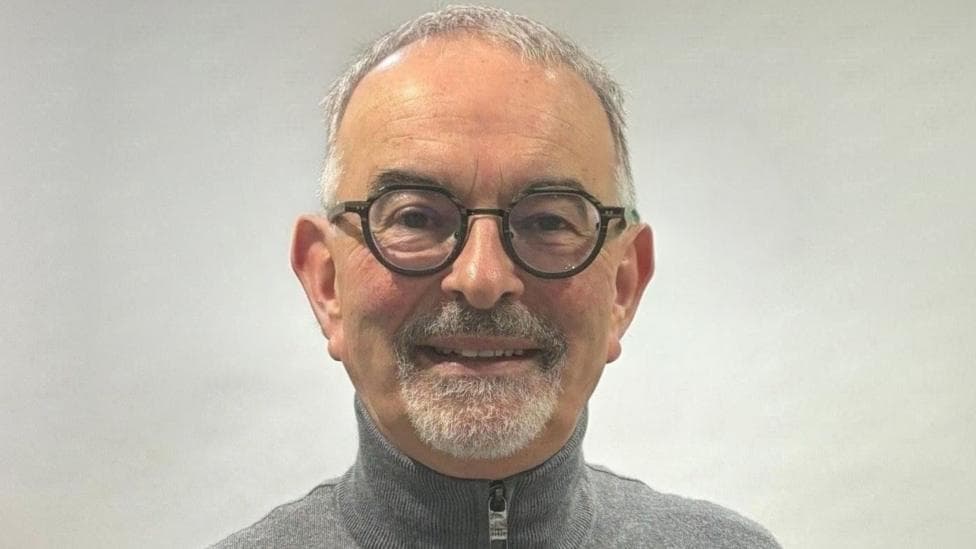 Dónal Doherty pictured wearing dark-rimmed glasses and in a grey zip-up sweater. He is standing in front of grey wall