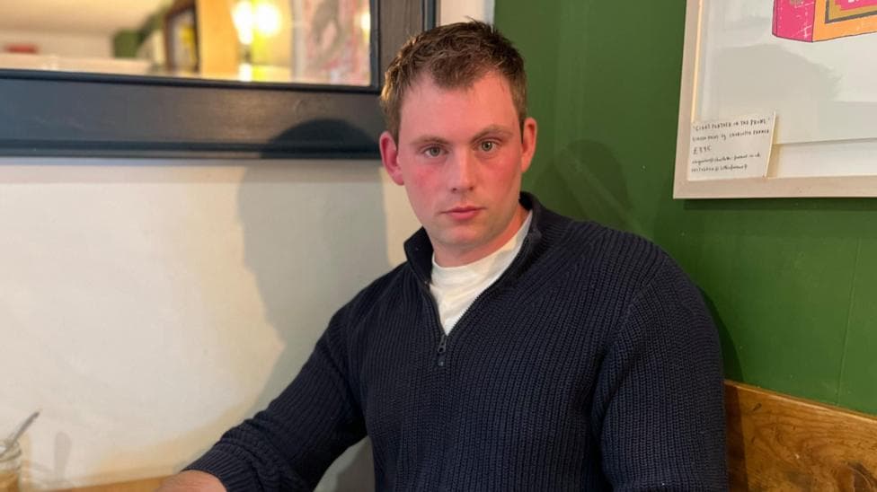 Callum is wearing a white t-shirt and blue jumper. He is looking directly at the camera.
