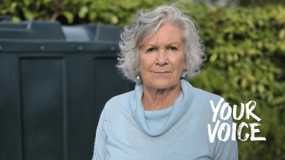 Roslynn Garner-Gatley has short, curly grey hair, is wearing a light blue high-collar jumper and round drop earrings, and has a small clip-on microphone attached near the neckline.
She is outside standing in front of her black heating oil tank