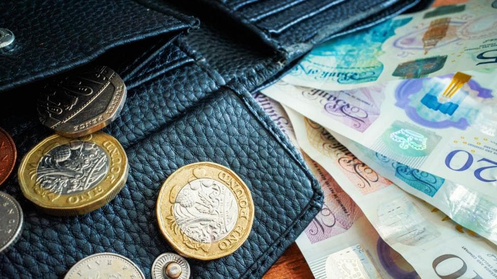 UK British pounds notes and coins lying beside a black wallet.