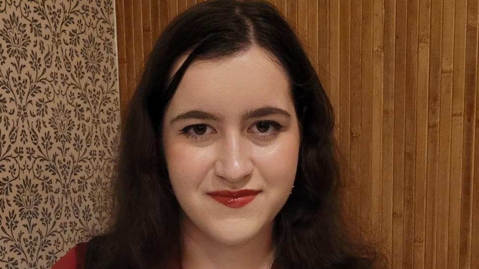 Carla looks at the camera. She has brown eyes and thick long dark hair. She is wearing red liptstick and there is a wooden wall behind her.