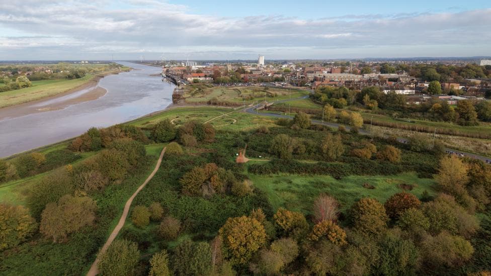 A drone shot of a large green area with lots of trees. A river runs down the left side of the green space.