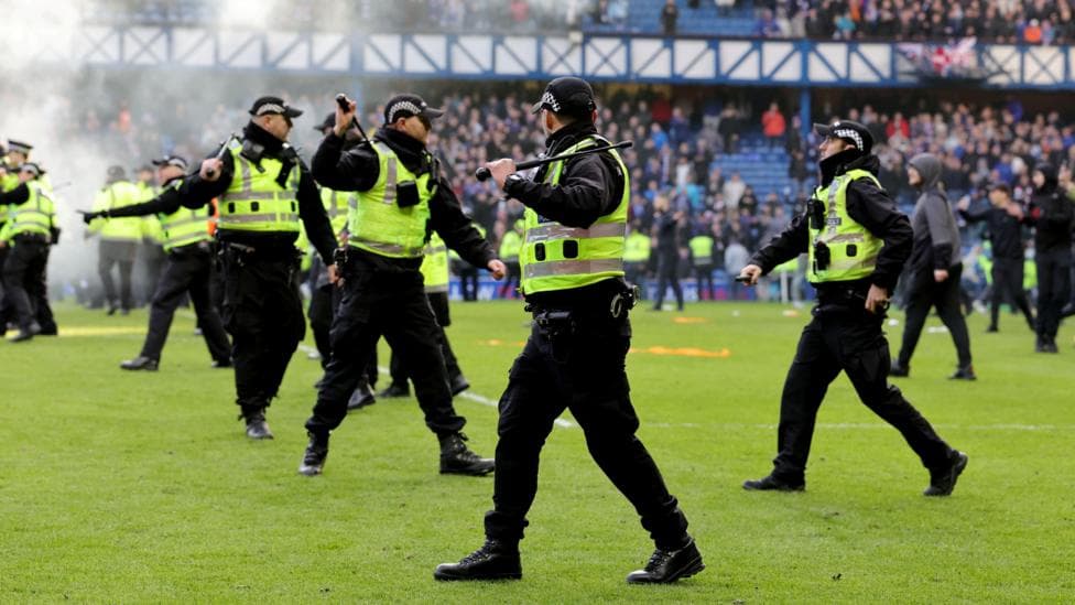 Police officers wearing black trousers, black jackets, hi-vis vests and black caps holding raised batons on a football pitch. There is smoke in the background. Fans can be seen on the pitch and in the stands.