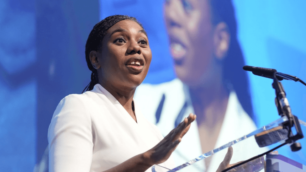Head and shoulders shot of Badenoch speaking a a glass podium. She is wearing a white top and has her black hair tied back in a low ponytail.