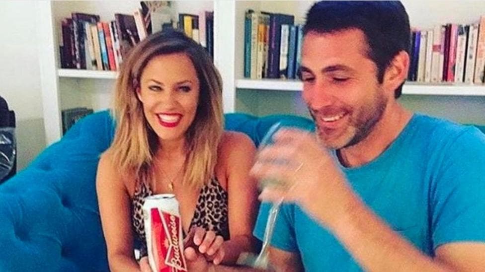 A woman smiles while looking towards a man with a blue T-shirt taking a drink from a glass while sat on a blue sofa in a room with books on a bookcase behind them.