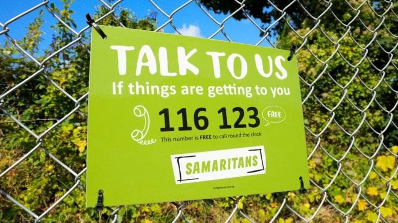 A banner for Samaritans charity attached to a wired fence.
It is green, white and black.
