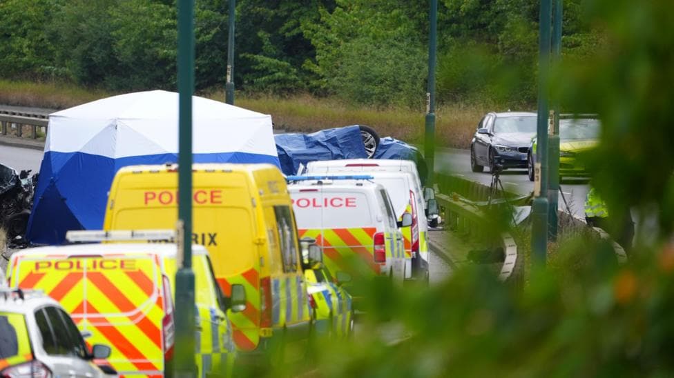 An overturned car is seen under a blue tarp, surrounded by eight police vehicles on a road. There are bushes in the background and street lights along the road kerb.