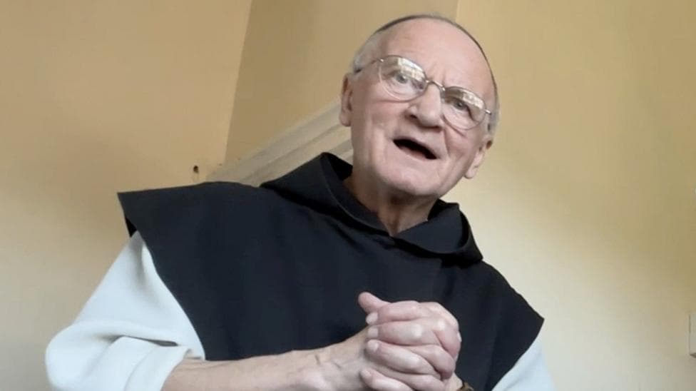 Brother Titus Keet is looking down at the photographer, with his hands clasped. He is wearing monks robes - a white tunic with a black scapular. He is open mouthed as if talking. He wears glasses and has very little hair.