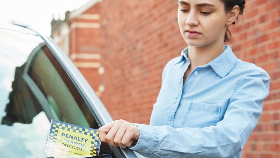 A woman in a blue shirt lifts a penalty charge notice from the front of her car