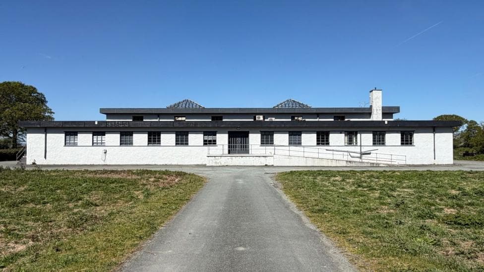 A long, white building at the end of a tarmac driveway with many windows and two protruding skylights.