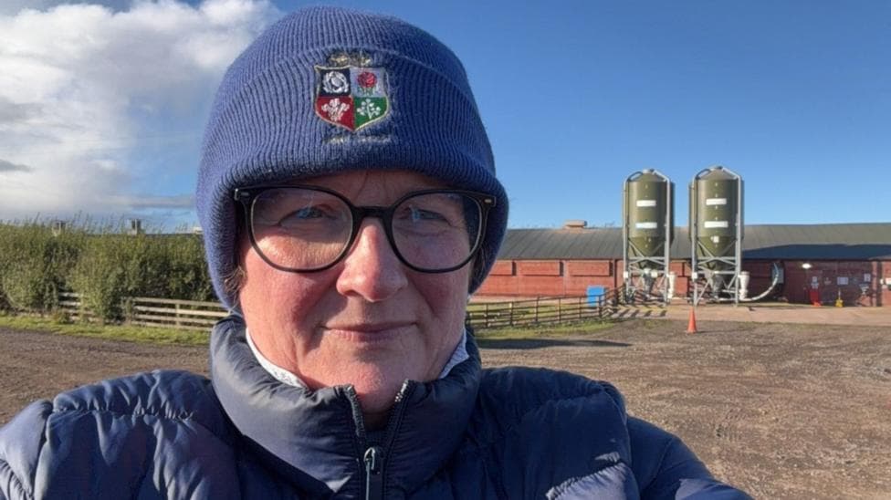 In a selfie-style head-and-shoulders shot, Catherine is wearing glasses, a blue woolly hat and matching puffer jacket. In the background is the farmyard and a long poultry barn.